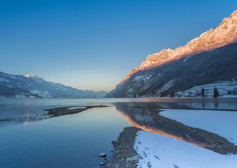 Morgenstimmung am winterlichen Walensee. Die Morgensonne strahlt die Spitzen der umliegende Berge in orangenem Licht an. Die Berge und die Wege um den See sind mit einer leichten Schneeschicht bedeckt.
