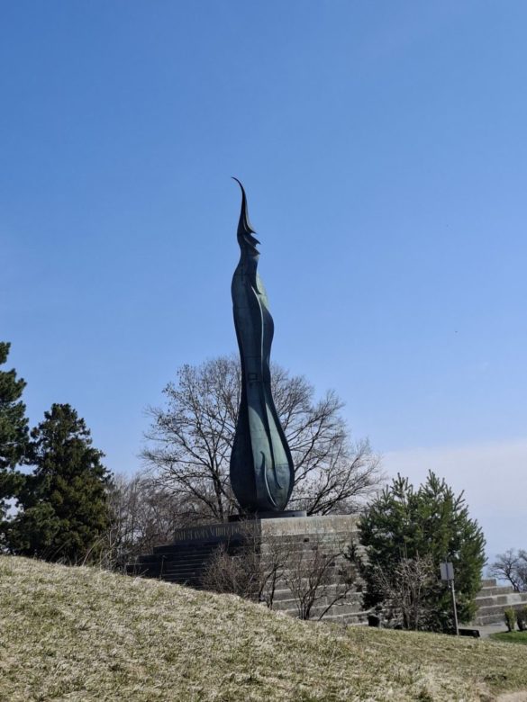 Das Foto zeigt eine hohe, schlanke Skulptur im Freien vor einem klaren, blauen Himmel. Die Skulptur steht auf einem gestuften Steinsockel auf einer Rasenfläche. Sie ist dunkel und metallisch, wirkt tropfen- oder flammenförmig und läuft nach oben in eine schmale, geschwungene Spitze aus. Im Hintergrund wachsen einige laublose Bäume sowie immergrüne Nadelbäume. Im Vordergrund ist leicht ansteigender, heller Rasen zu sehen, der noch winterlich oder frühjährlich wirkt. Die Szene vermittelt den Eindruck eines Aussichtspunktes oder Denkmals in einer offenen Parklandschaft.