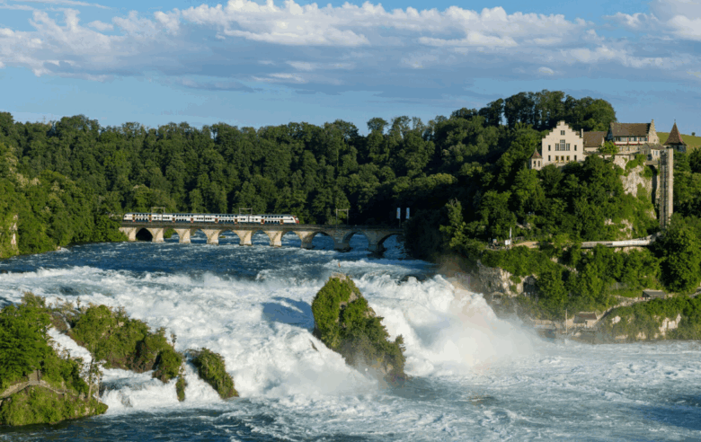 Ein breiter, wuchtiger Wasserfall des Rheins stürzt über mehrere Felsstufen hinab, im Vordergrund wirbeln weiße Wassermassen, links führen Wege mit Geländern an den Felsen entlang, im Hintergrund überquert ein Zug auf einer steinernen Bogenbrücke den Fluss und rechts oben thront Schloss Laufen mit hellen Gebäuden auf einem bewaldeten Hügel unter blauem Himmel.