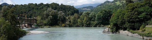 Panoramaaufnahme des Zusammenflusses von Vorder- und Hinterrhein bei Reichenau in Graubünden: Im Vordergrund fliesst ein breiter, hell türkisgrauer Fluss durch das Bild, links und rechts gesäumt von grünen Bäumen und Sträuchern, am linken Ufer sind eine Brücke und einzelne Gebäude zu erkennen, im Hintergrund erheben sich sanfte, teils bewaldete Hügel und dahinter höher gelegene Berge unter einem leicht bewölkten Himmel