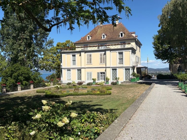 Das Foto zeigt das Schloss Arenenberg an einem sonnigen Tag. Das zweistöckige, hellgelbe Gebäude im französischen Stil besitzt ein braunrotes Walmdach mit Gauben. Davor liegen eine gepflegte Rasenfläche mit Blumenbeeten und ein Kiesweg. Im linken Hintergrund ist zwischen grünen Bäumen der Bodensee unter blauem Himmel zu erkennen.