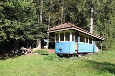 Ein kleiner, blau‑cremefarbener Eisenbahnwagen steht als umgebauter Aufenthaltsraum in einer Waldlichtung. Der Wagen hat ein dunkles Ziegeldach und mehrere Fenster entlang der Seite. Vor ihm steht ein Picknicktisch, an dem einige Menschen sitzen. Rundherum wachsen hohe Bäume, und die Szene wirkt ruhig und naturnah.